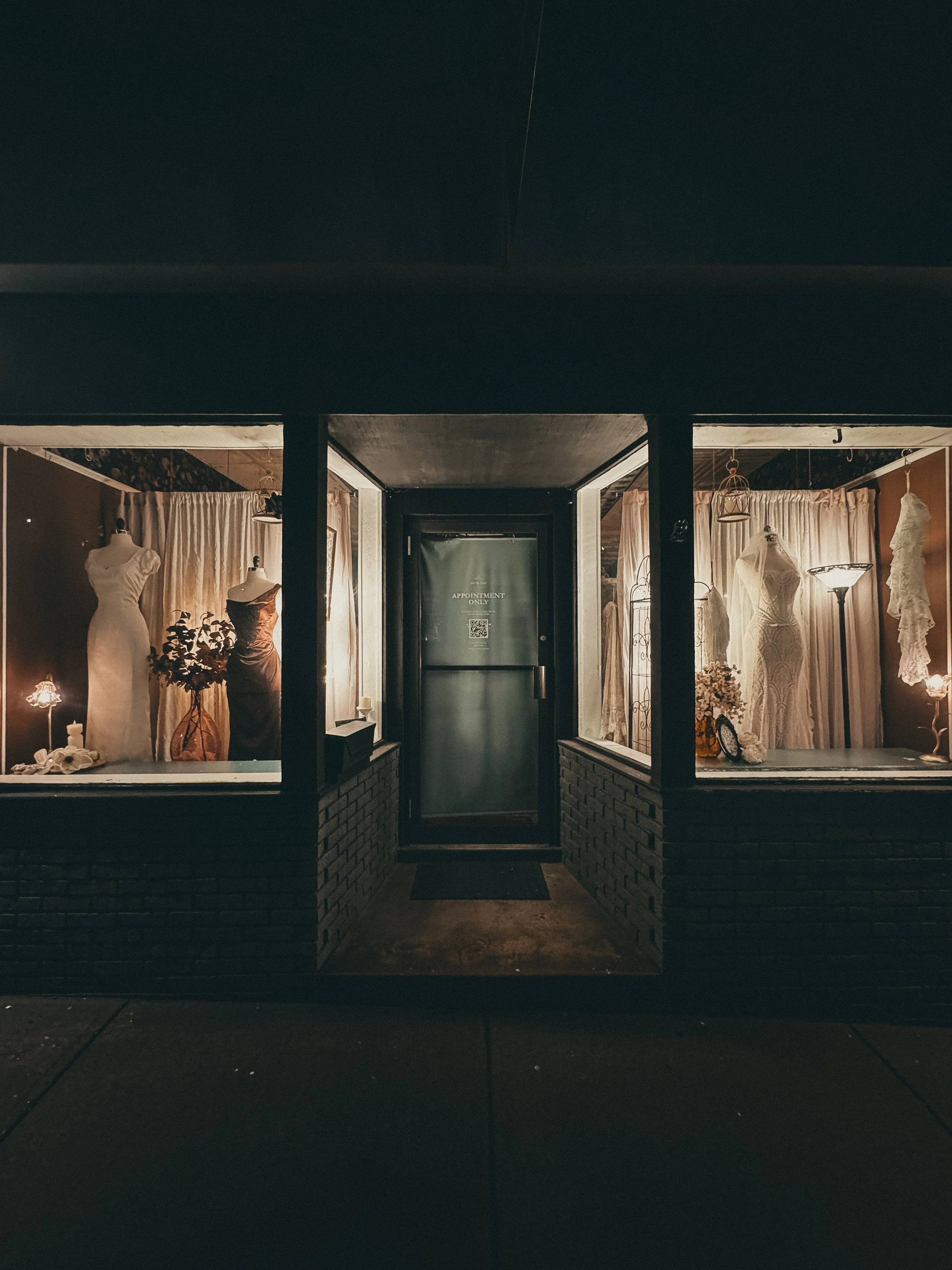 Nighttime view of a boutique bridal shop window displaying wedding dresses and mannequins, with warm lighting and curtains inside.