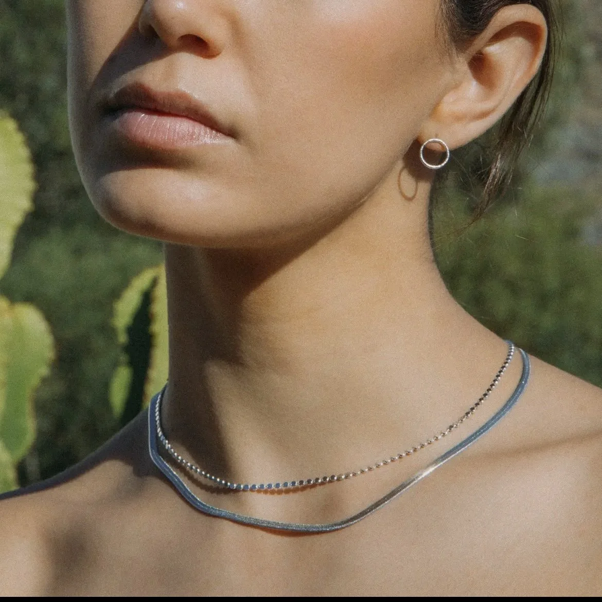 The Anatole Choker layered with a silver herringbone chain against a green leafy backdrop