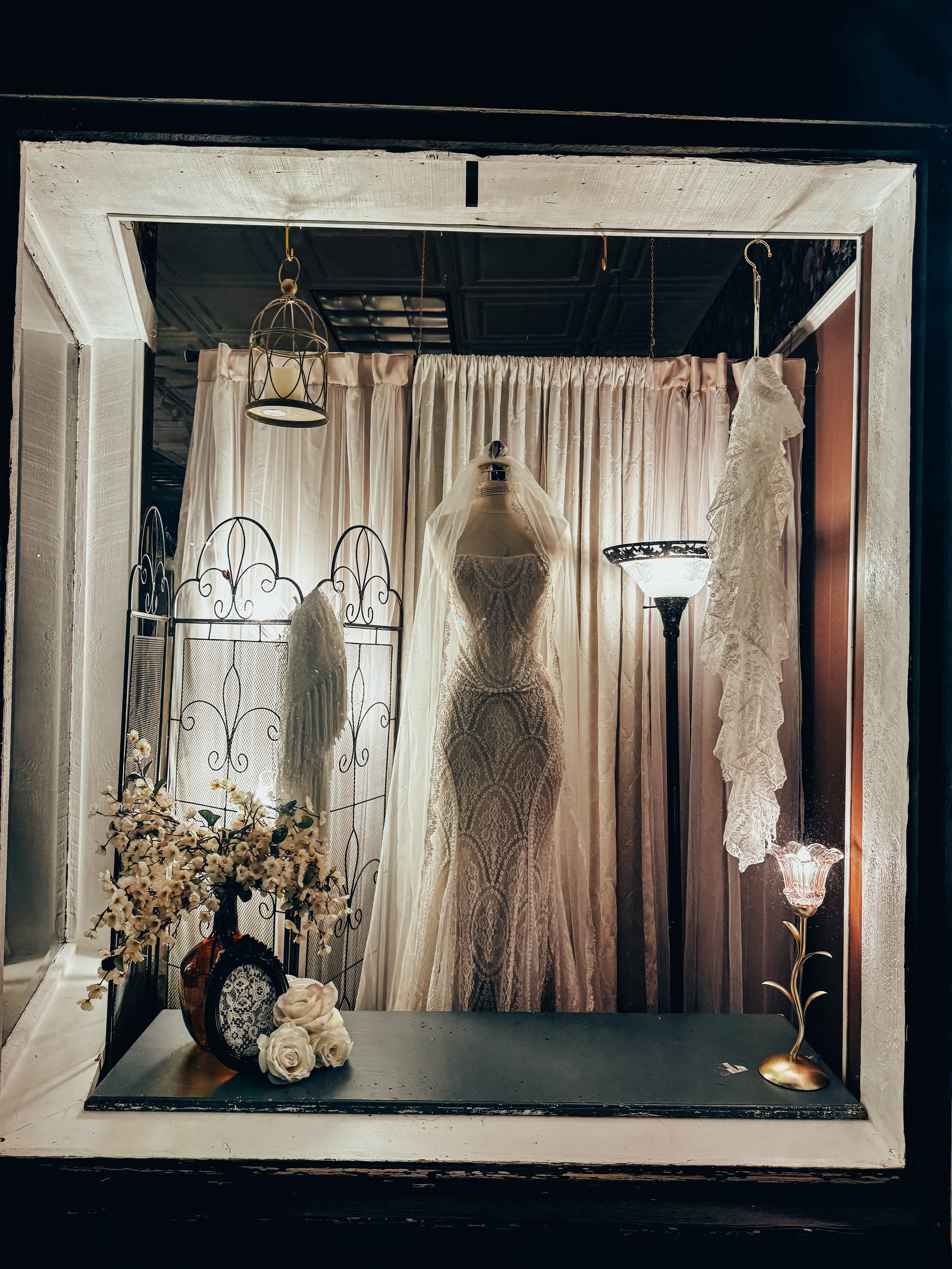 Display window with wedding dress on mannequin, floral arrangements, and vintage lamps.