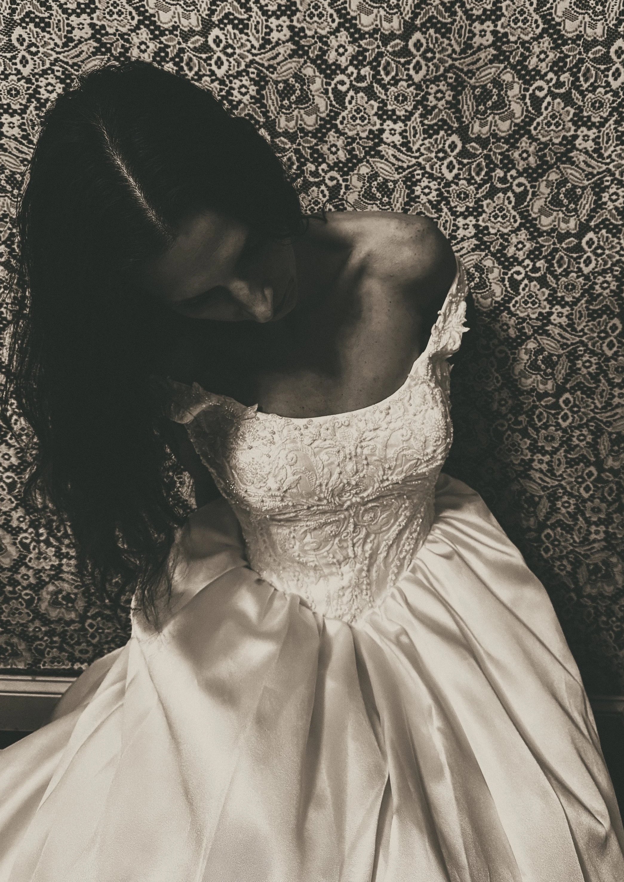 A black-and-white photo of a woman in a voluminous vintage wedding gown, leaning forward with her head tilted down. The lace backdrop frames her figure, creating an evocative, melancholic bridal portrait.