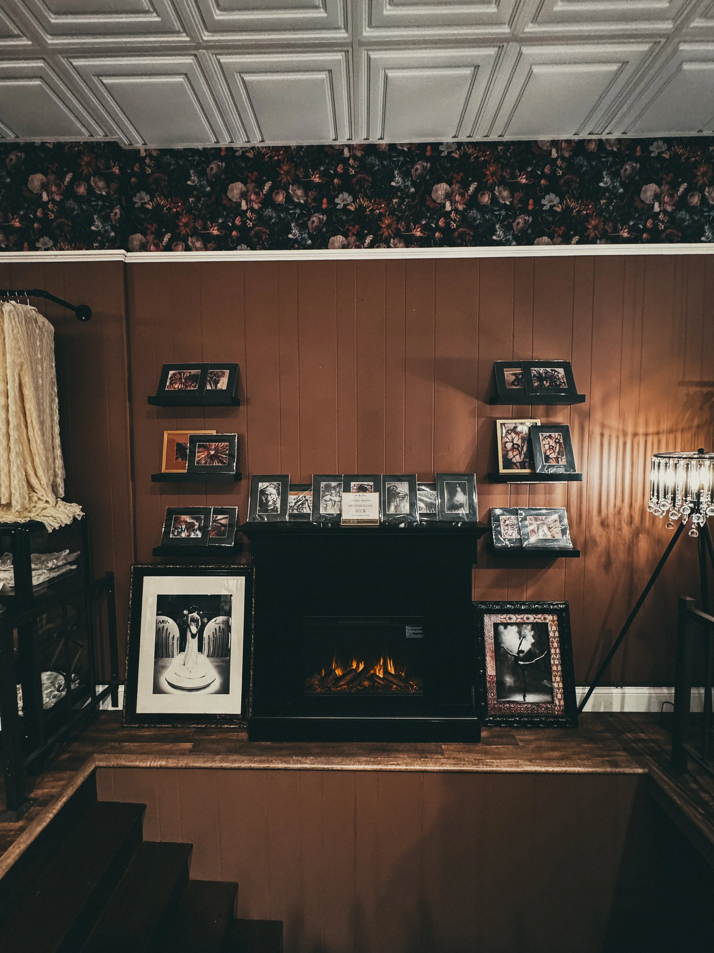 Photo of a wall with framed photographs and artwork surrounding a black electric fireplace. The wall has a wood panel design, with a floral wallpaper border at the top. There is a small table or stand on the left with folded fabrics or linens, and a 