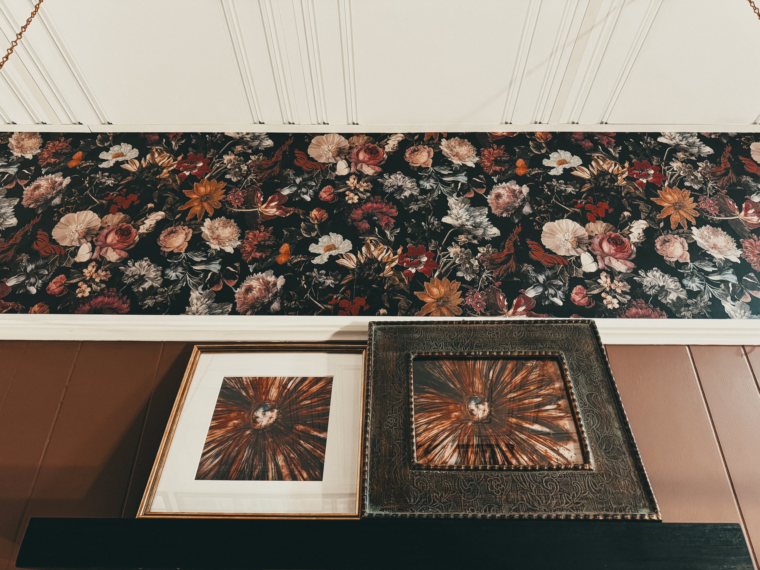 Decorative wall with floral wallpaper, framed artwork, and a decorative metal tray with an abstract design.