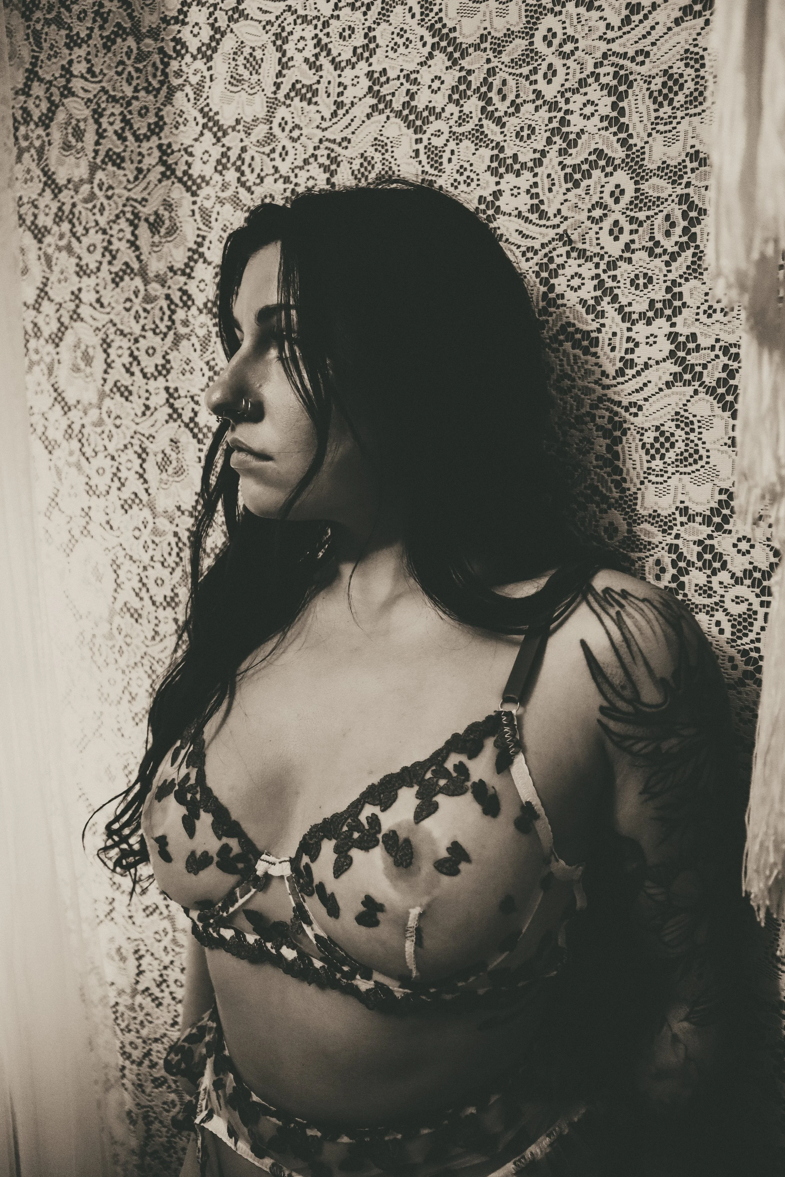 A woman with dark hair, tattoos, and a nose piercing, wearing a floral lingerie top, leaning against a lace curtain background.