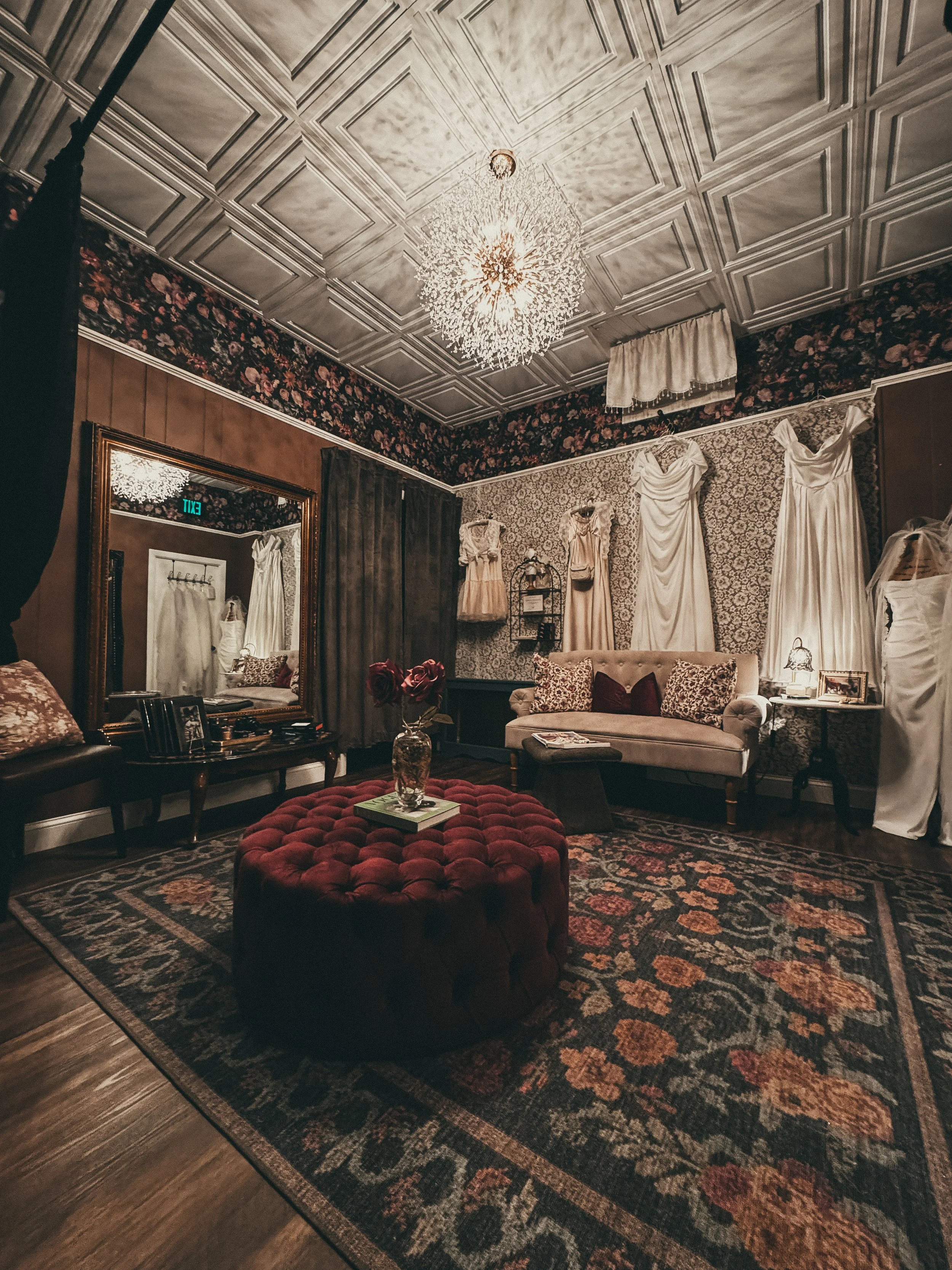A room with floral wallpaper, ornate ceiling tiles, a chandelier, a mirror, a sofa with patterned pillows, draped curtains, a red tufted ottoman, and a picture frame on a side table.