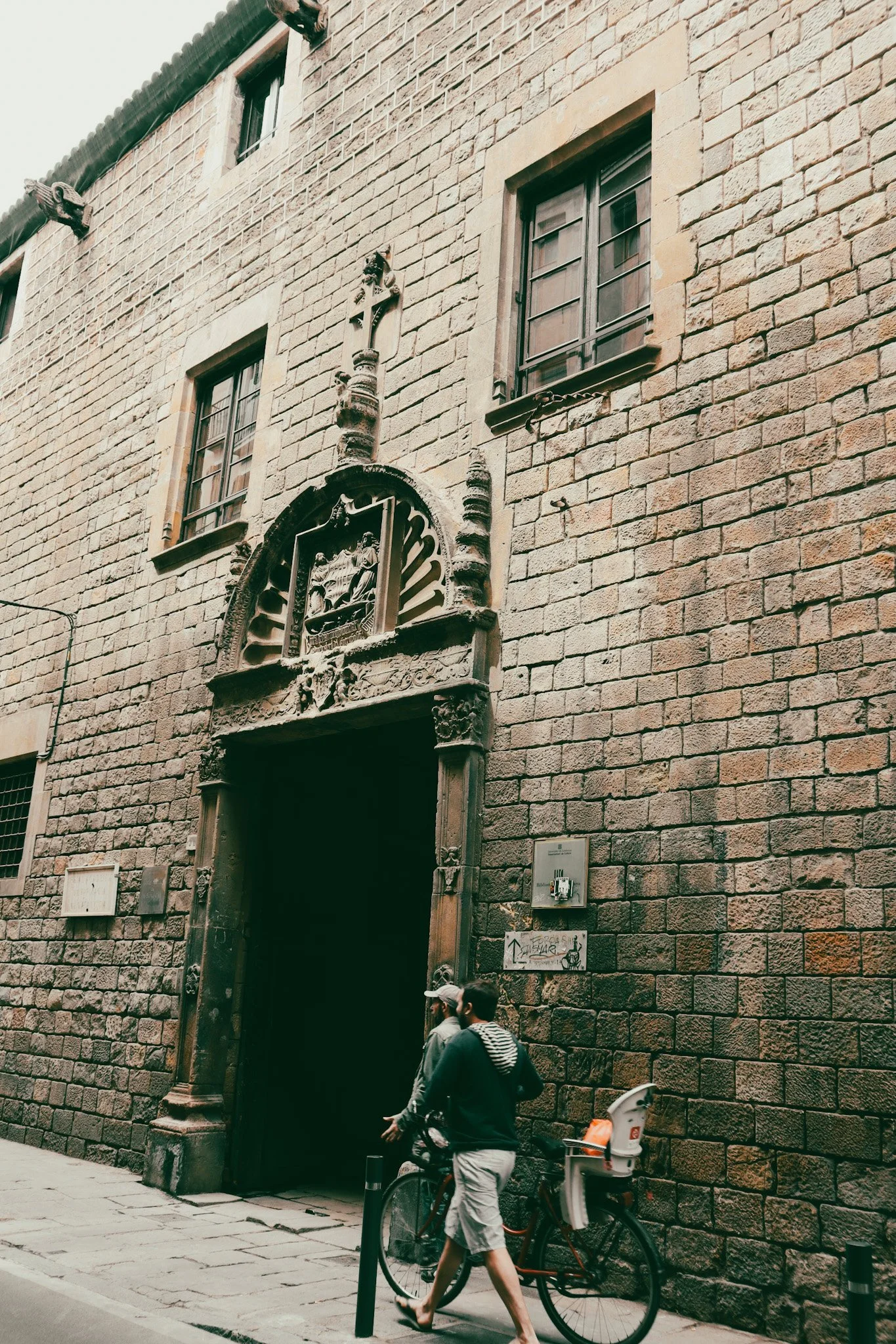 A historic brick building with an ornate religious sculpture above the entrance, featuring a small arched doorway, two windows above, and pedestrians and a bicycle nearby.