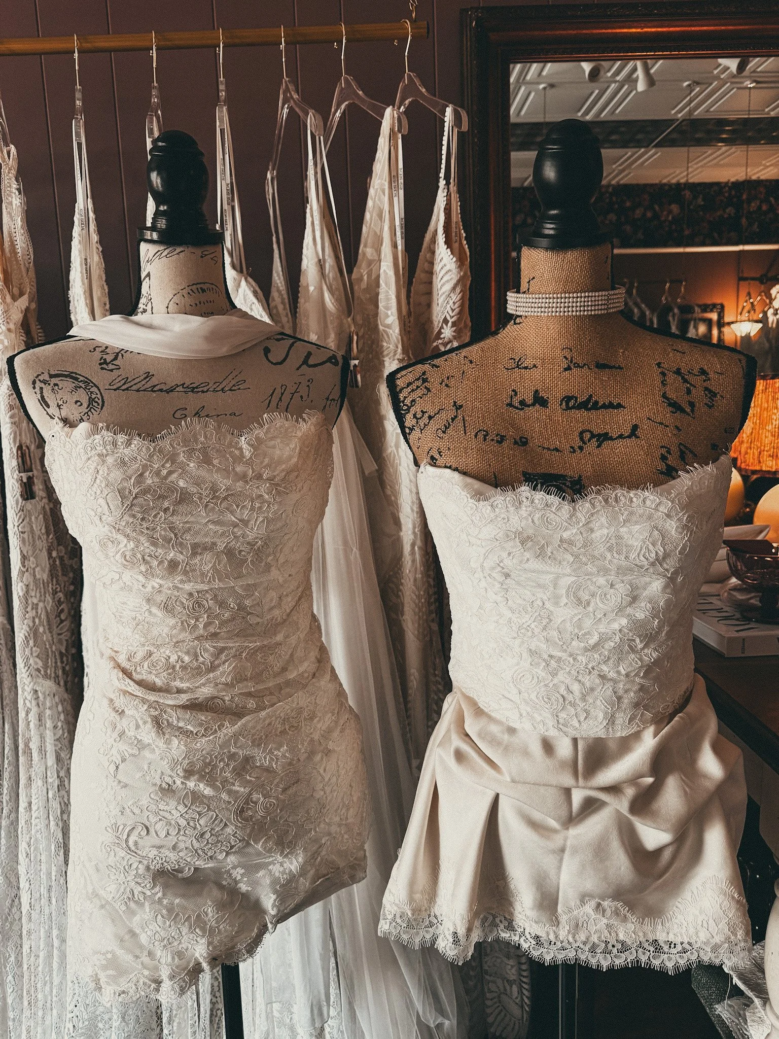 Two dress mannequins displaying vintage-style strapless wedding dresses, with lace detailing, against a backdrop of hanging lace fabrics and a mirror.