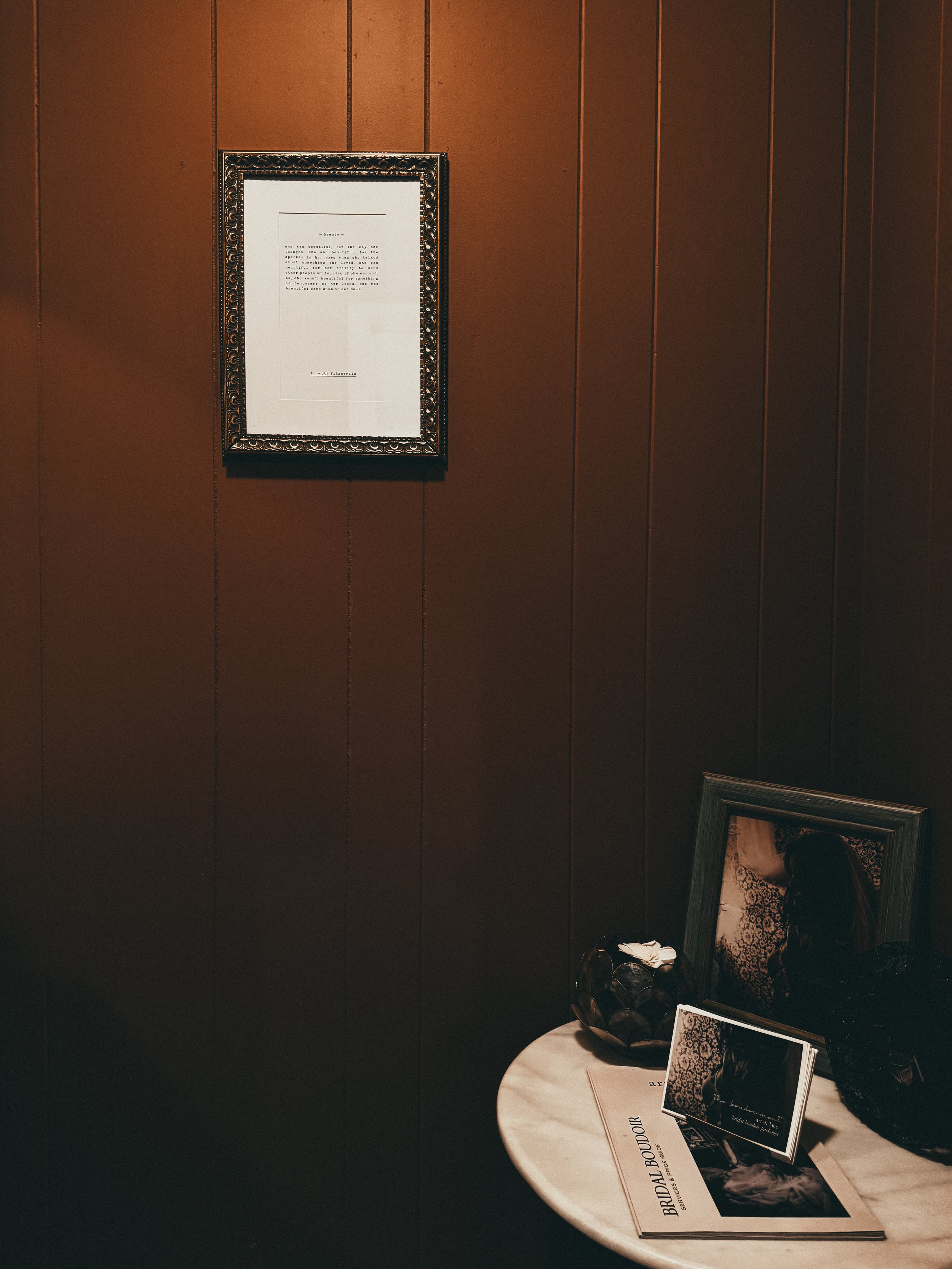 A corner of a room with dark wood paneled wall, decorated with framed photographs and artwork on a small round table. The table holds magazines, a framed picture, and decorative objects. The framed photograph depicts a woman with long hair.