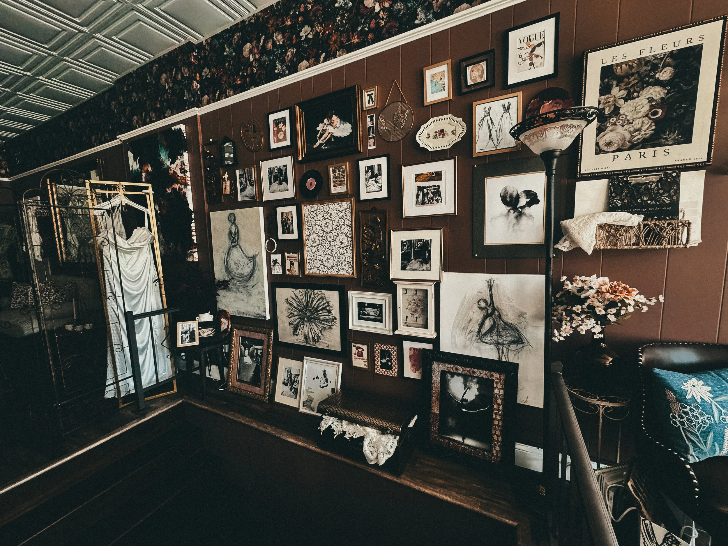 A wall with a collage of various framed photos, artwork, and prints in a cozy, vintage-style room. The wall features a lamp, floral decoration, and a clothing rack on the left, with a dark wood bench beneath the framed art. The room has a dark wood-paneled wall, floral wallpaper at the top, and a patterned ceiling.