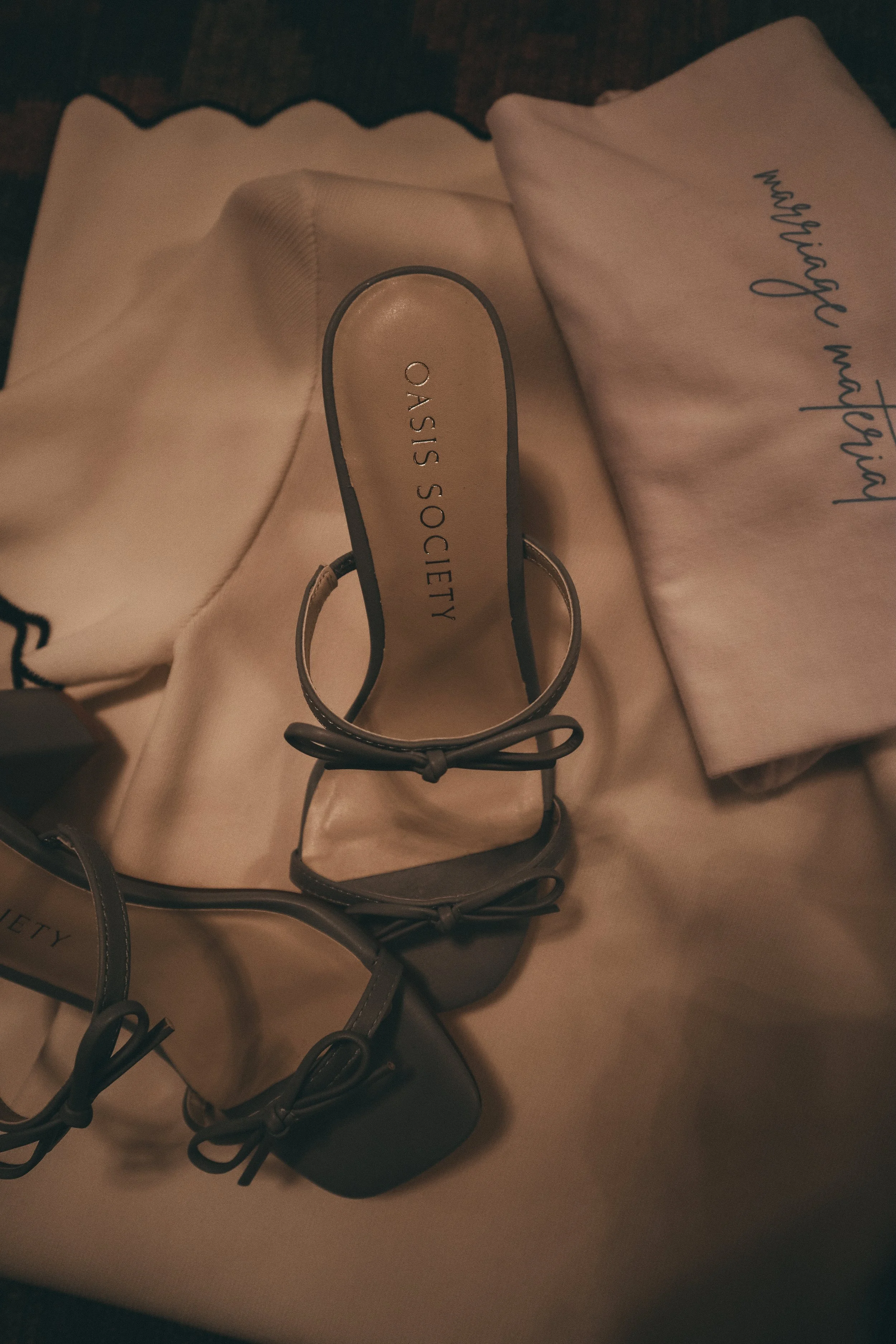 A beige high-heeled shoe with an ankle strap and bow detail from Oasis Society on a beige cloth surface. There is also a folded piece of pale pink fabric with handwritten words.