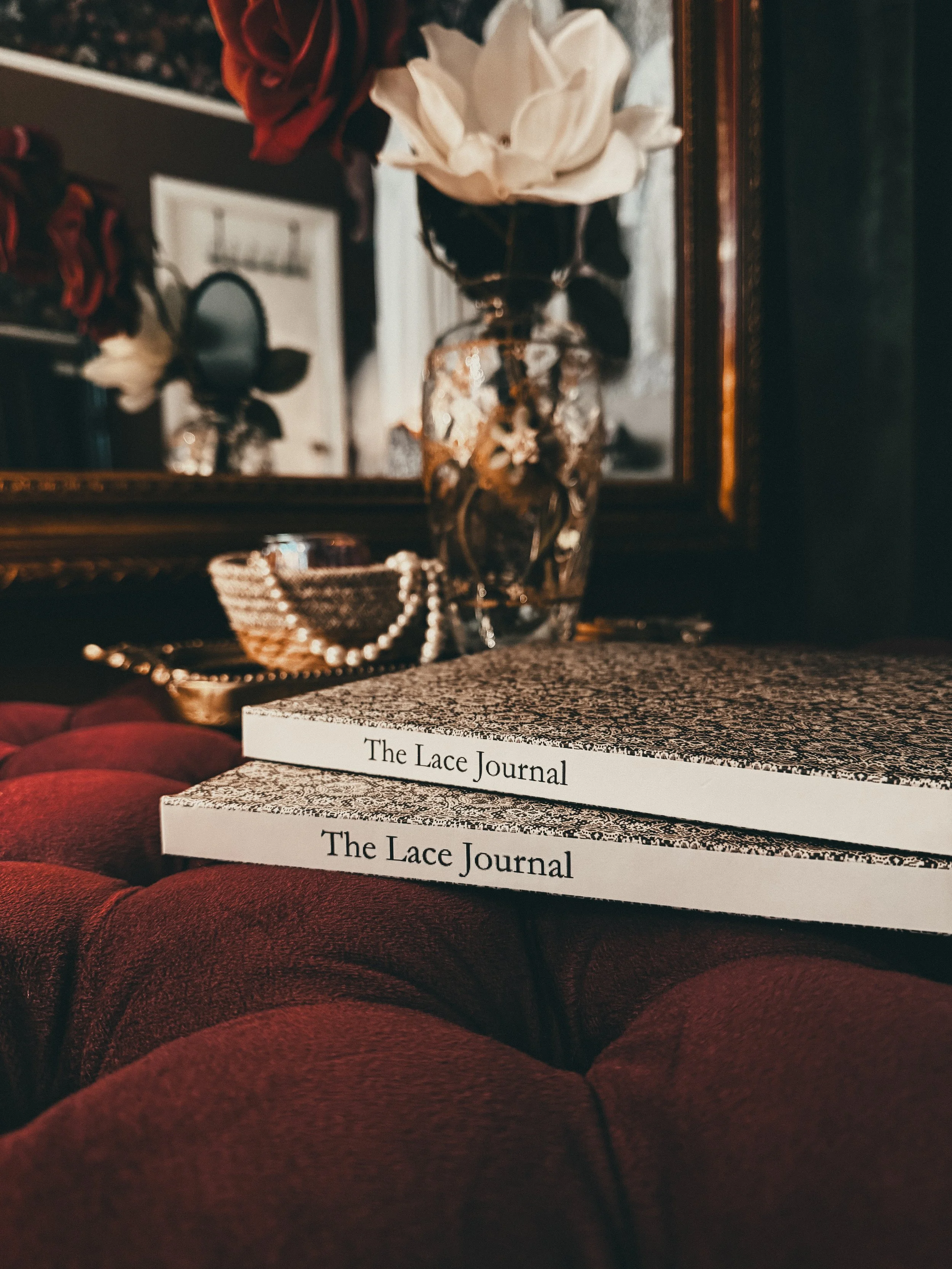 Two stacked books titled 'The Lace Journal' on a red, tufted surface with a glass vase holding a white flower and dark foliage, a jewelry box with pearls, and a mirror with a blurred reflection in the background.