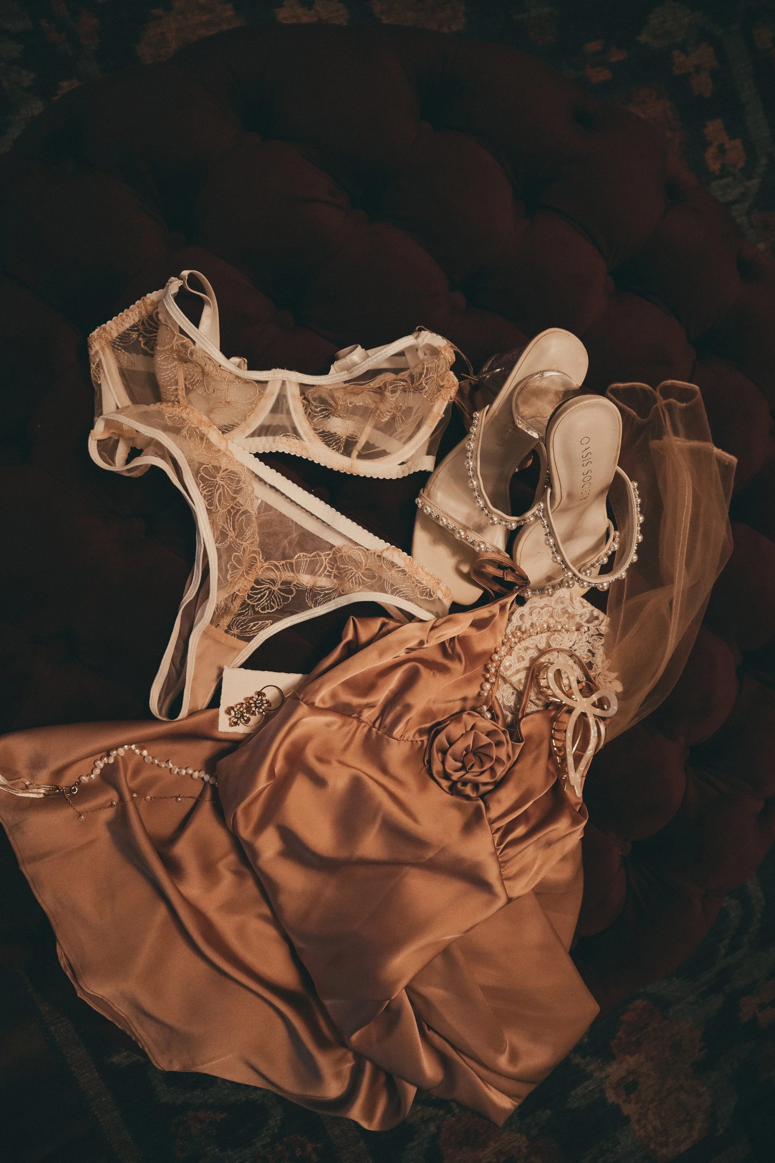Lingerie, shoes, jewelry, and a satin dress laid out on a dark tufted surface, possibly for an elegant occasion.