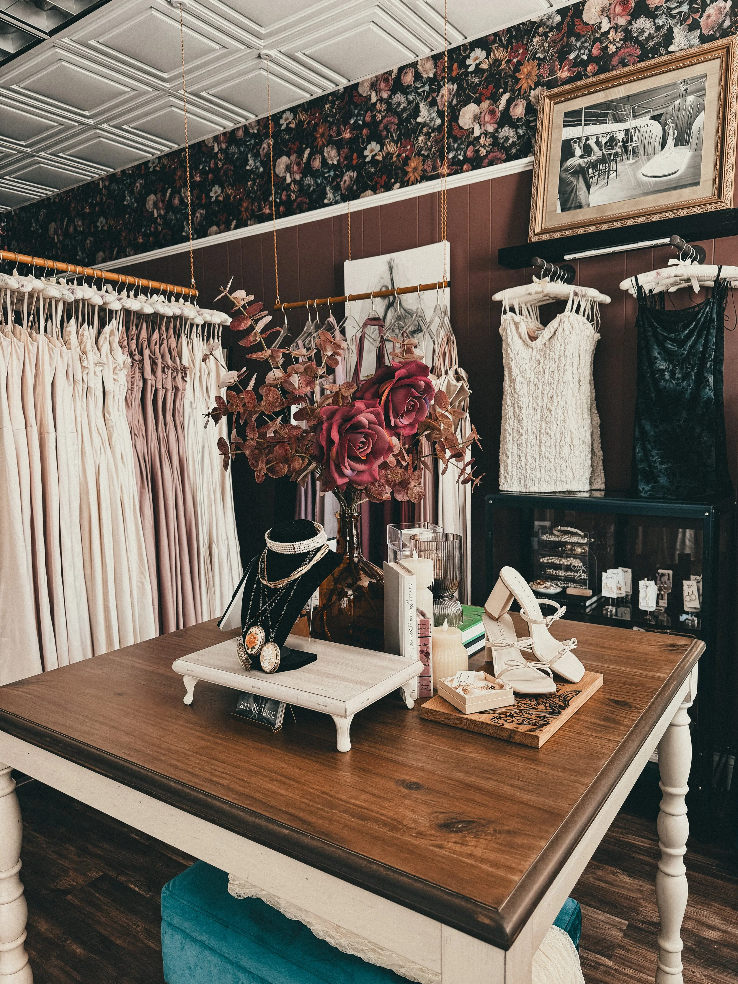 Interior of a boutique with dresses hanging on racks, floral wallpaper, framed pictures on the wall, and a wooden table displaying jewelry, shoes, books, and a large vase with pink roses.
