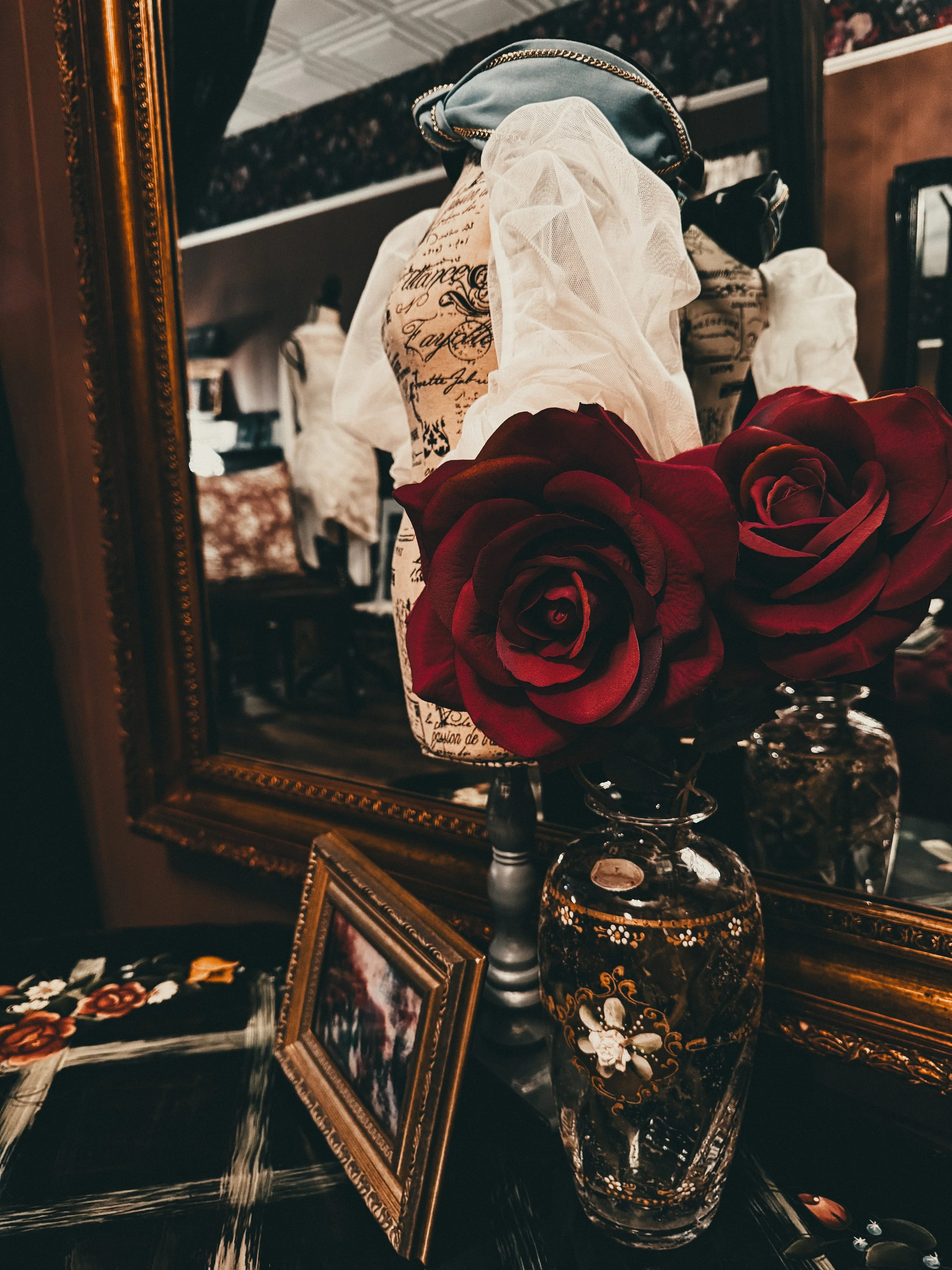 A vintage dresser with a large ornate mirror, a glass vase with red roses, a small framed photograph, and decorative jewelry or trinkets, all reflected in the mirror.