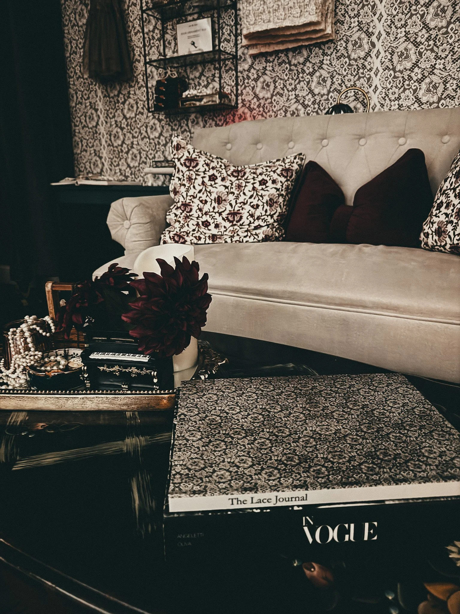 A beige upholstered sofa with floral pillows, a black jewelry box filled with jewelry, a plant with dark red leaves, a magazine titled The Lace Journal, and a black Vogue magazine. The background features patterned wallpaper with black and beige orna