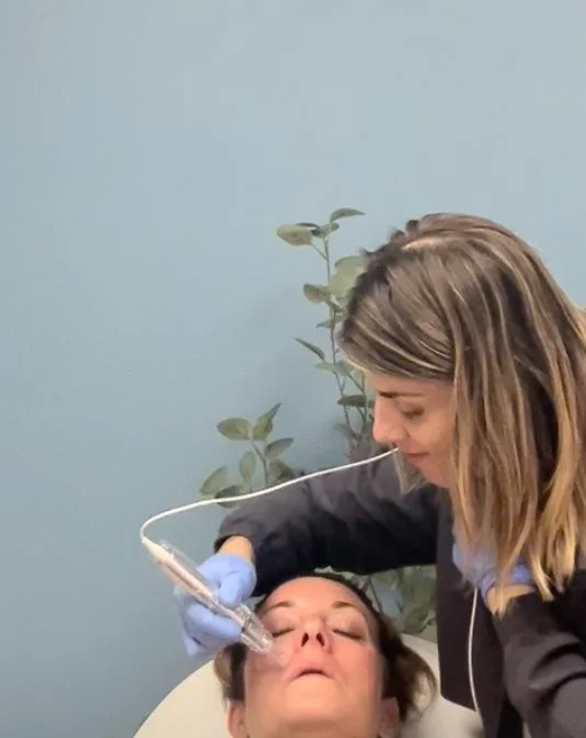 A woman undergoing a facial treatment performed by a professional using a handheld device, wearing gloves, in a spa or clinic setting.