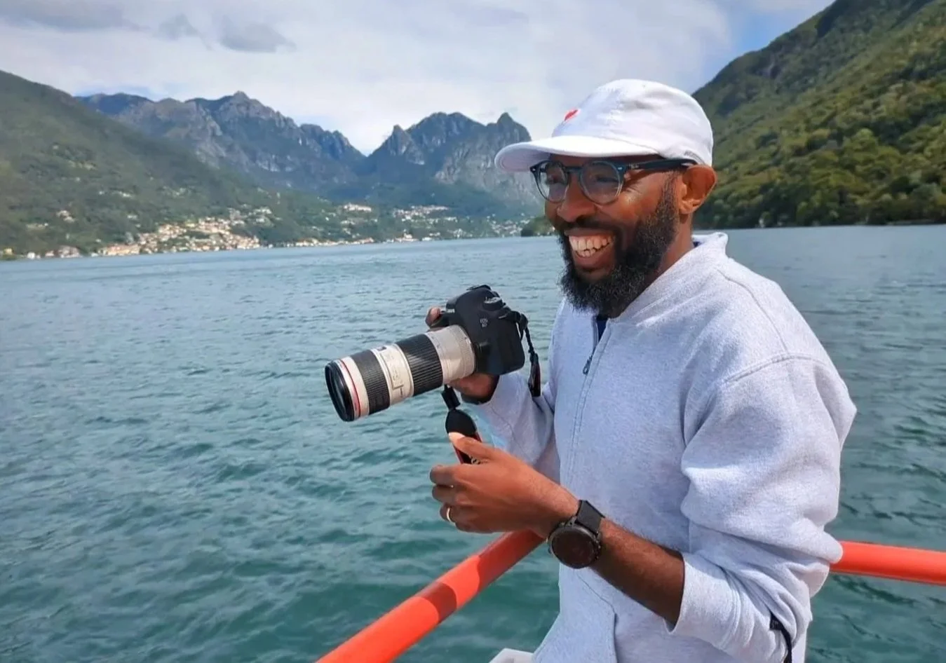 A man with glasses and a beard aboard a boat holding a camera with a large telephoto lens, smiling, with a lake and mountains in the background.
