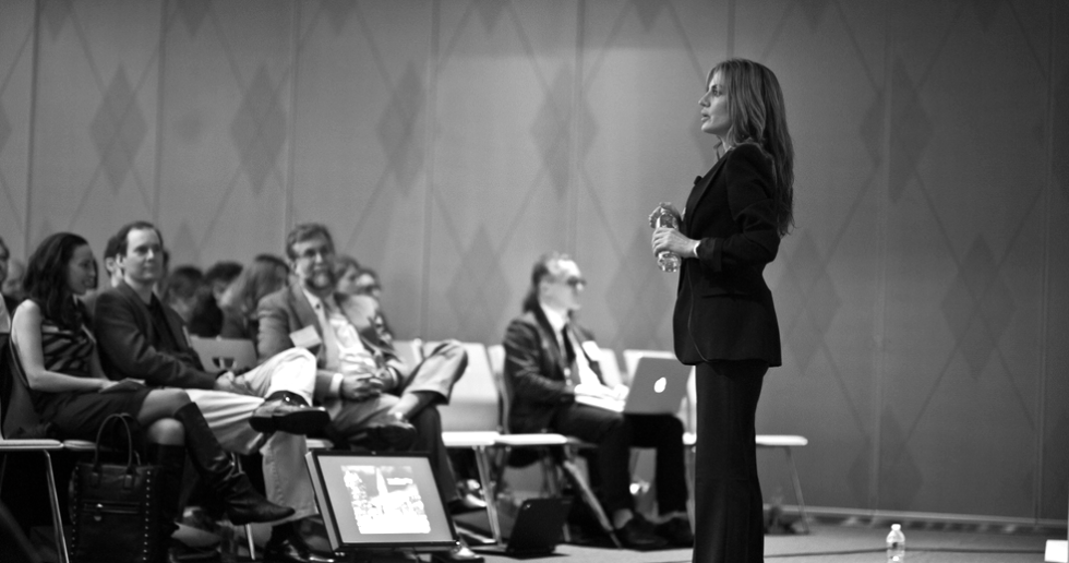 A woman in black standing and speaking to an audience in a conference room.