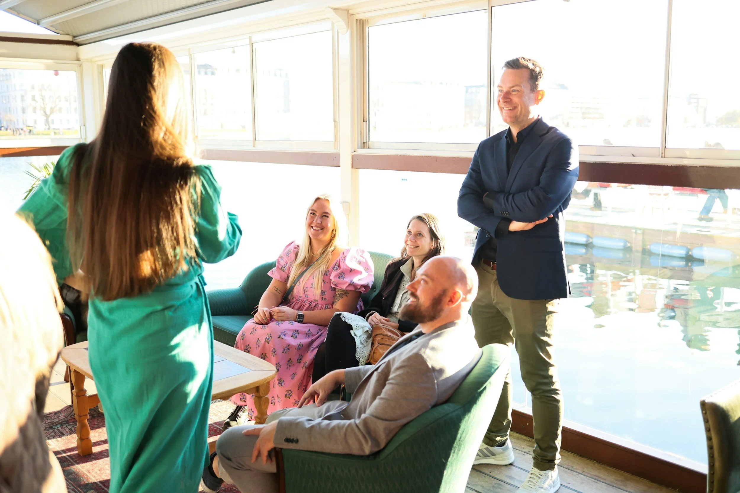 Group of people in a well-lit room, with some sitting on sofas and others standing, engaged in conversation and smiling, with large windows in the background showing a waterfront view.