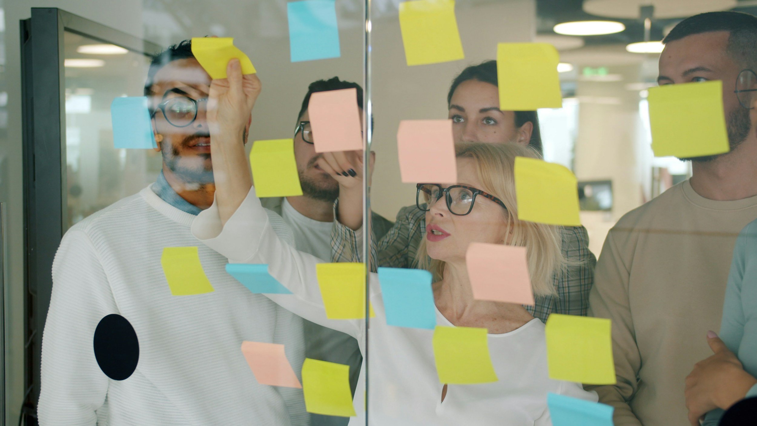 Group of five diverse coworkers brainstorming and discussing ideas, with colorful sticky notes on a glass wall in an office.