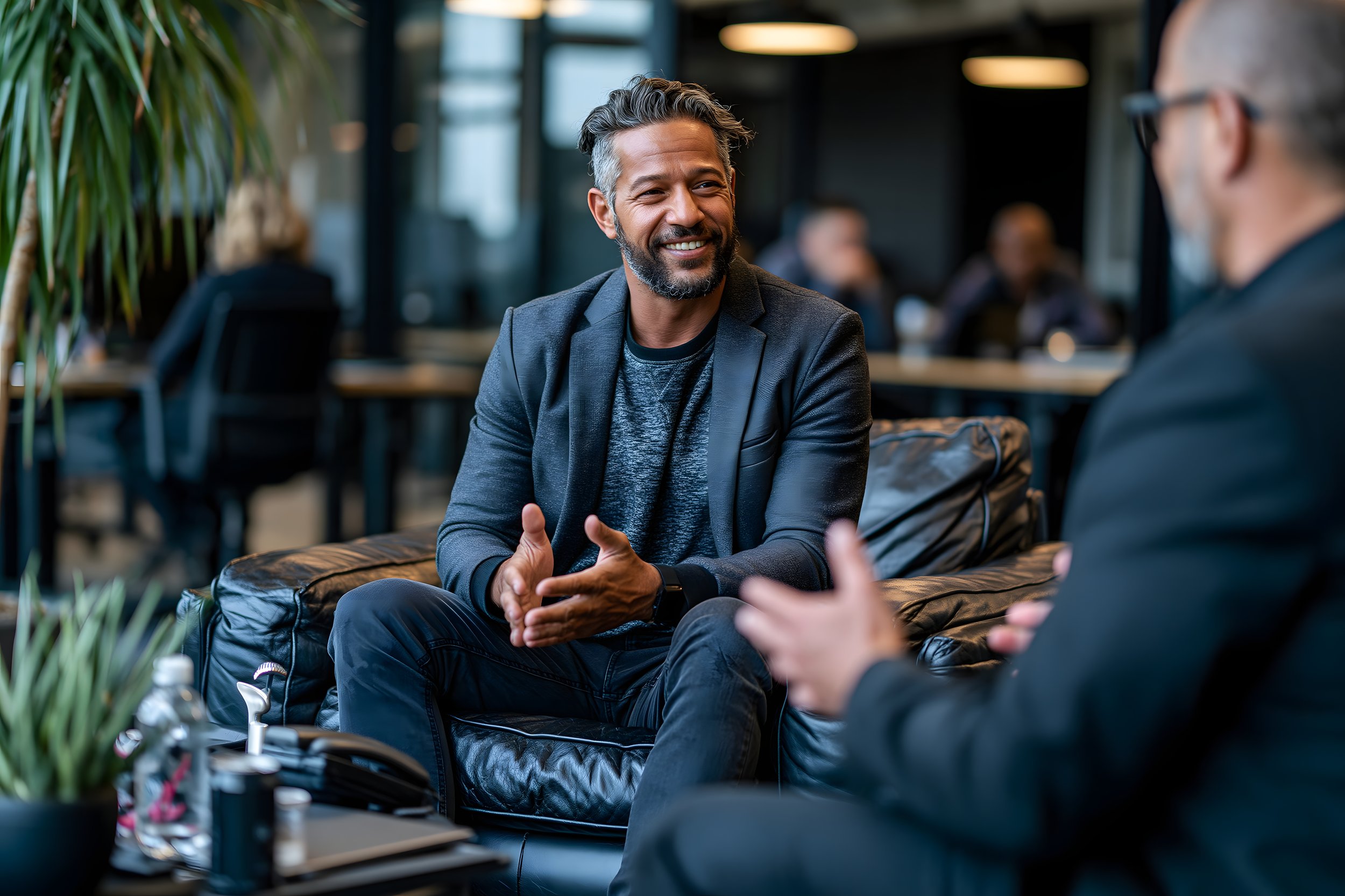 A middle-aged man with salt-and-pepper hair and beard, wearing a dark blazer and shirt, smiling and talking to another person in a modern office or cafe setting, with other people in the background.