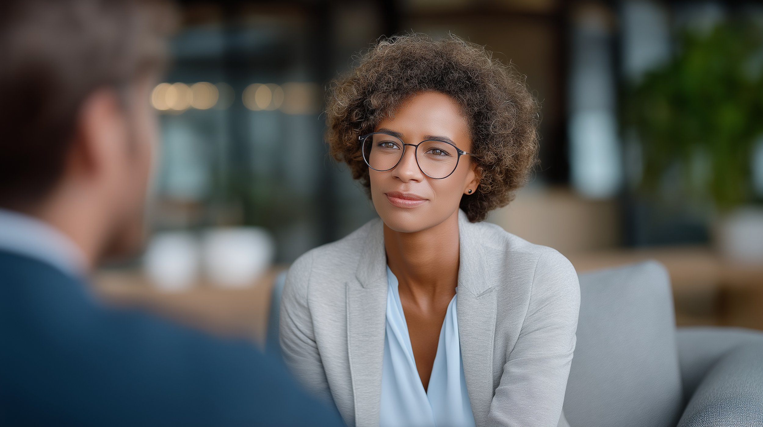 A woman with curly hair and glasses interviewed or having conversation with a man, with a blurred background.
