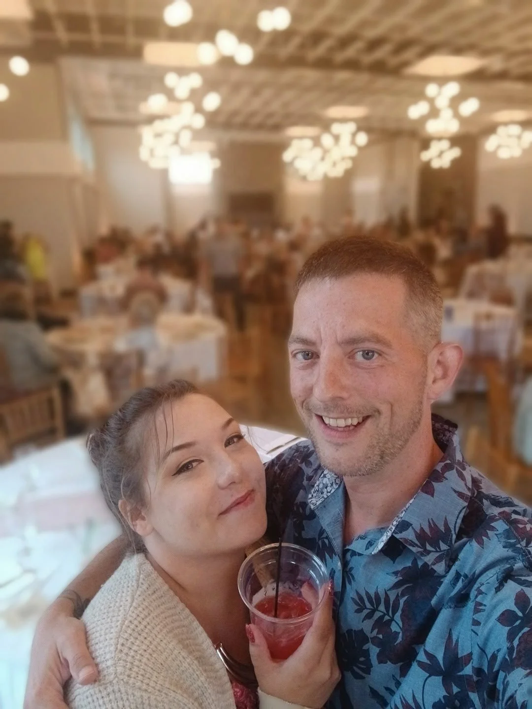 A man and woman taking a selfie at a crowded indoor event hall, with the woman holding a red drink in a clear plastic cup.