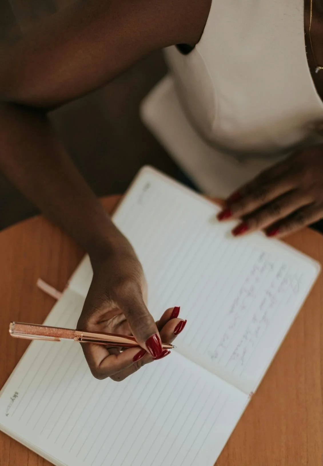 A person with dark skin and red painted nails writing in a lined notebook with a pink pen. The person is wearing a white sleeveless top and has a gold bracelet on their wrist.
