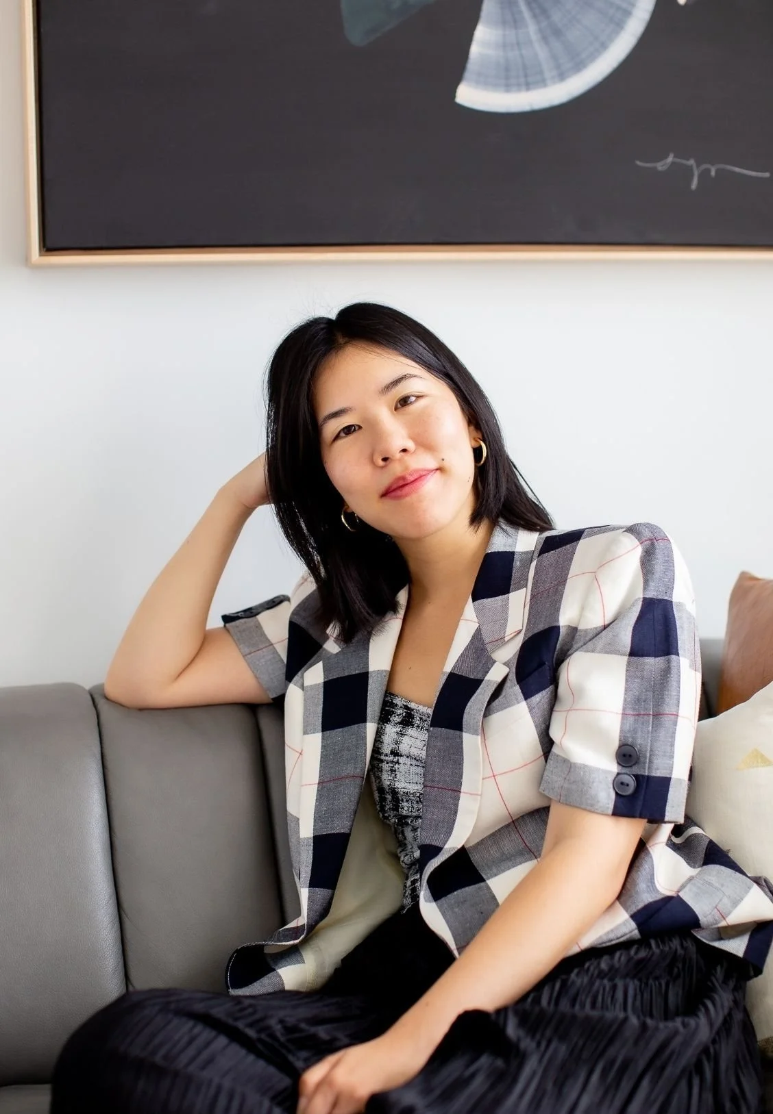 Shawn Li, Online Therapist in New York City, with shoulder-length black hair sitting on a gray sofa, wearing a black and white checkered blazer, smiling at the camera with her right hand resting behind her head, in a room with art hanging behind her.