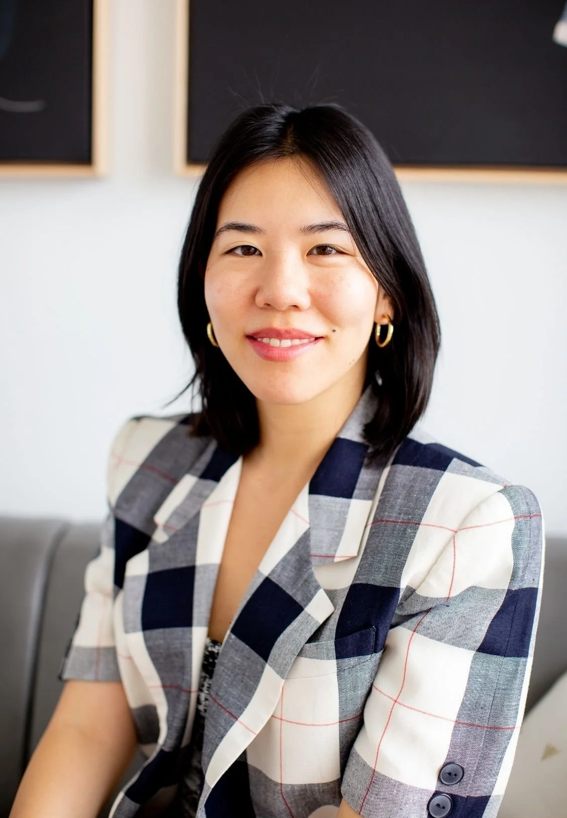 Shawn Li, Online Therapist in New York City, with shoulder-length black hair, wearing gold hoop earrings and a plaid blazer, smiling at the camera in an indoor setting.