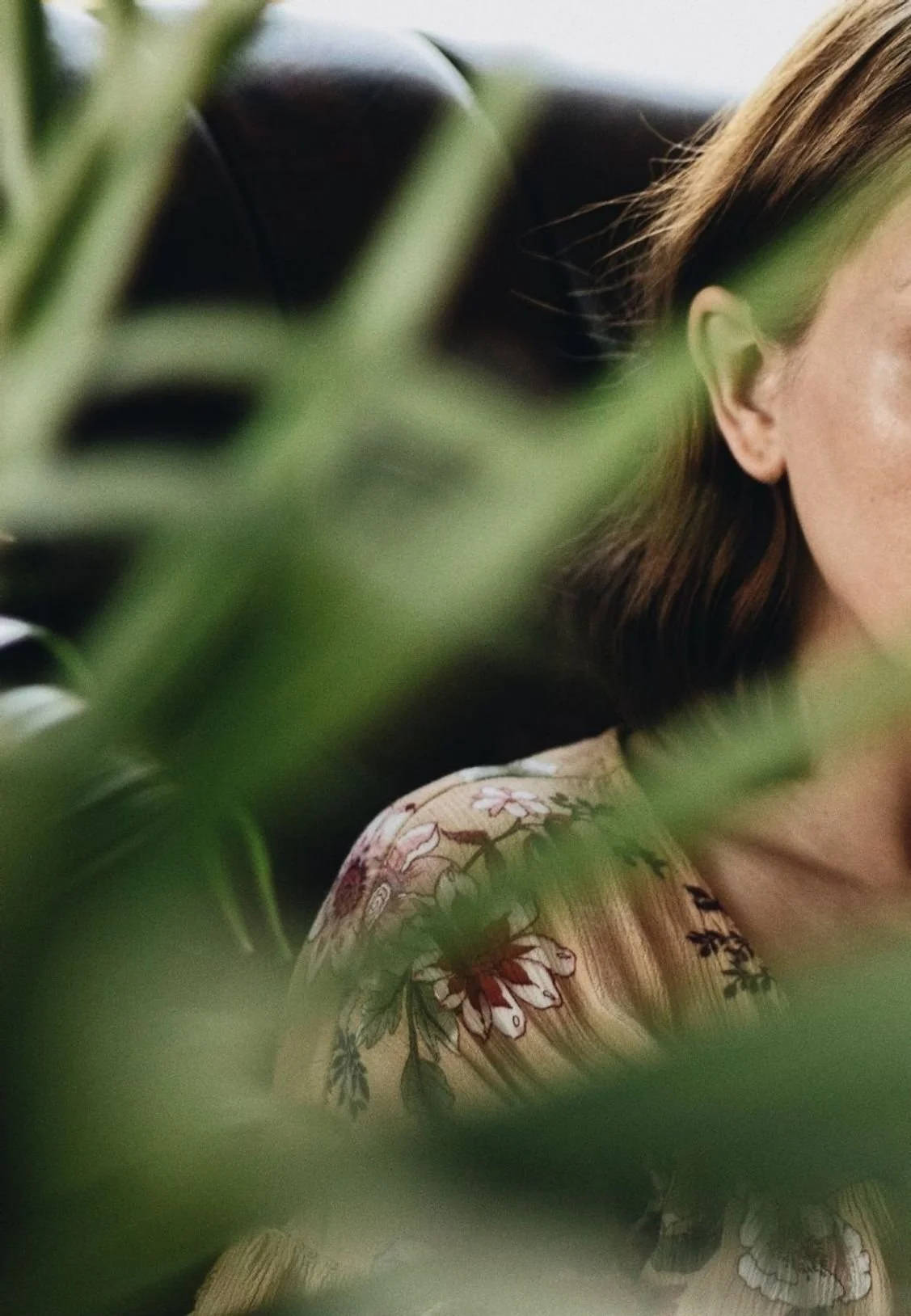Close-up of a woman with brown hair, seen through green leaves, wearing a floral-patterned garment.