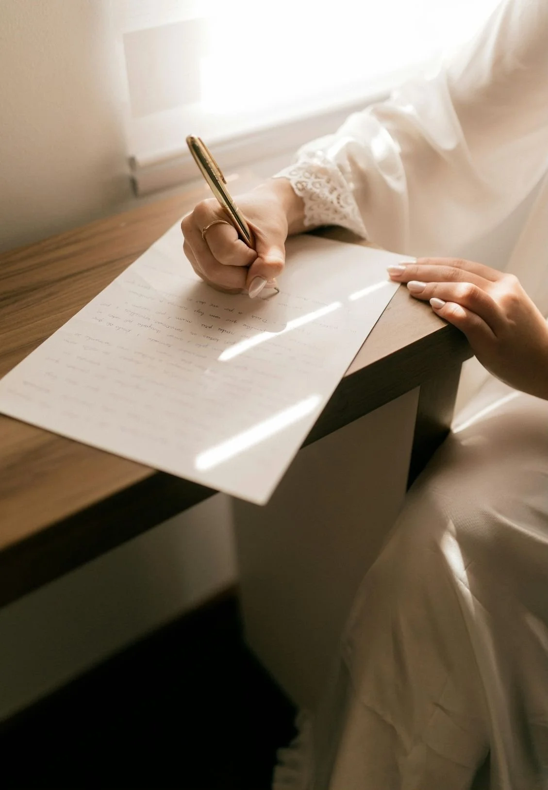 A person wearing a long, white, lace-cuffed sleeve is writing on a piece of paper with a gold pen, sitting at a wooden desk near a window with sunlight coming through.