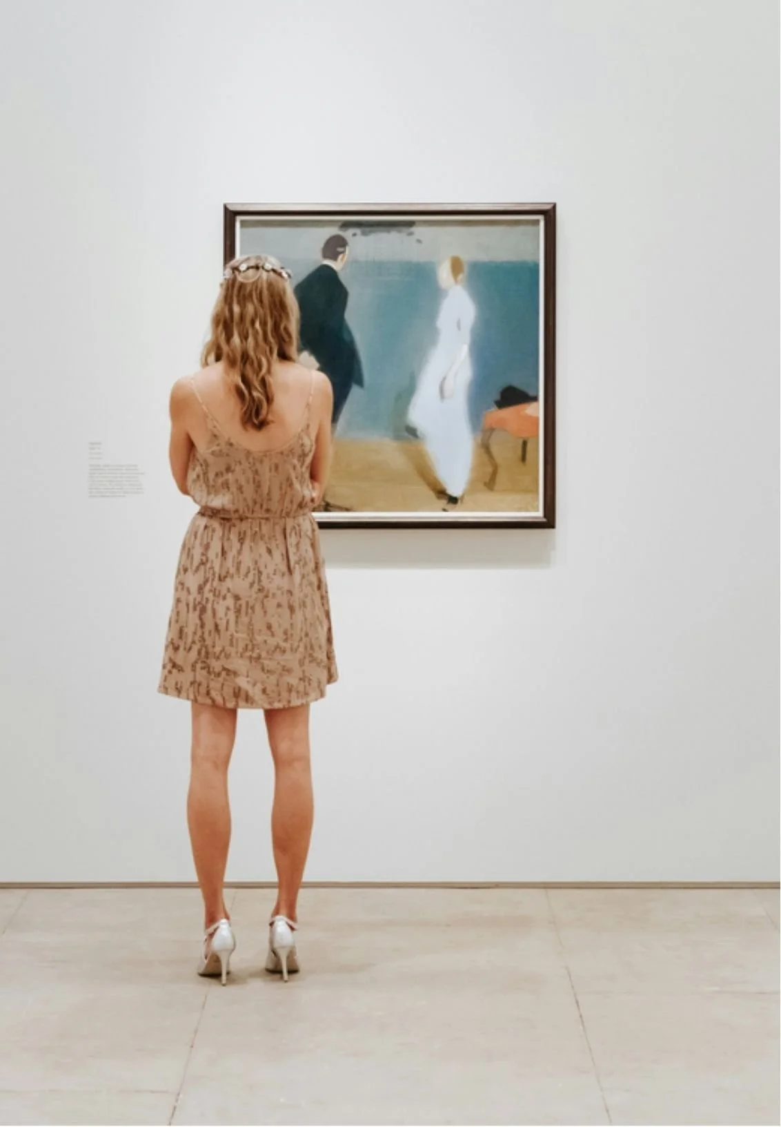A woman in a light brown, patterned dress and white heels stands in front of an art painting at a museum.