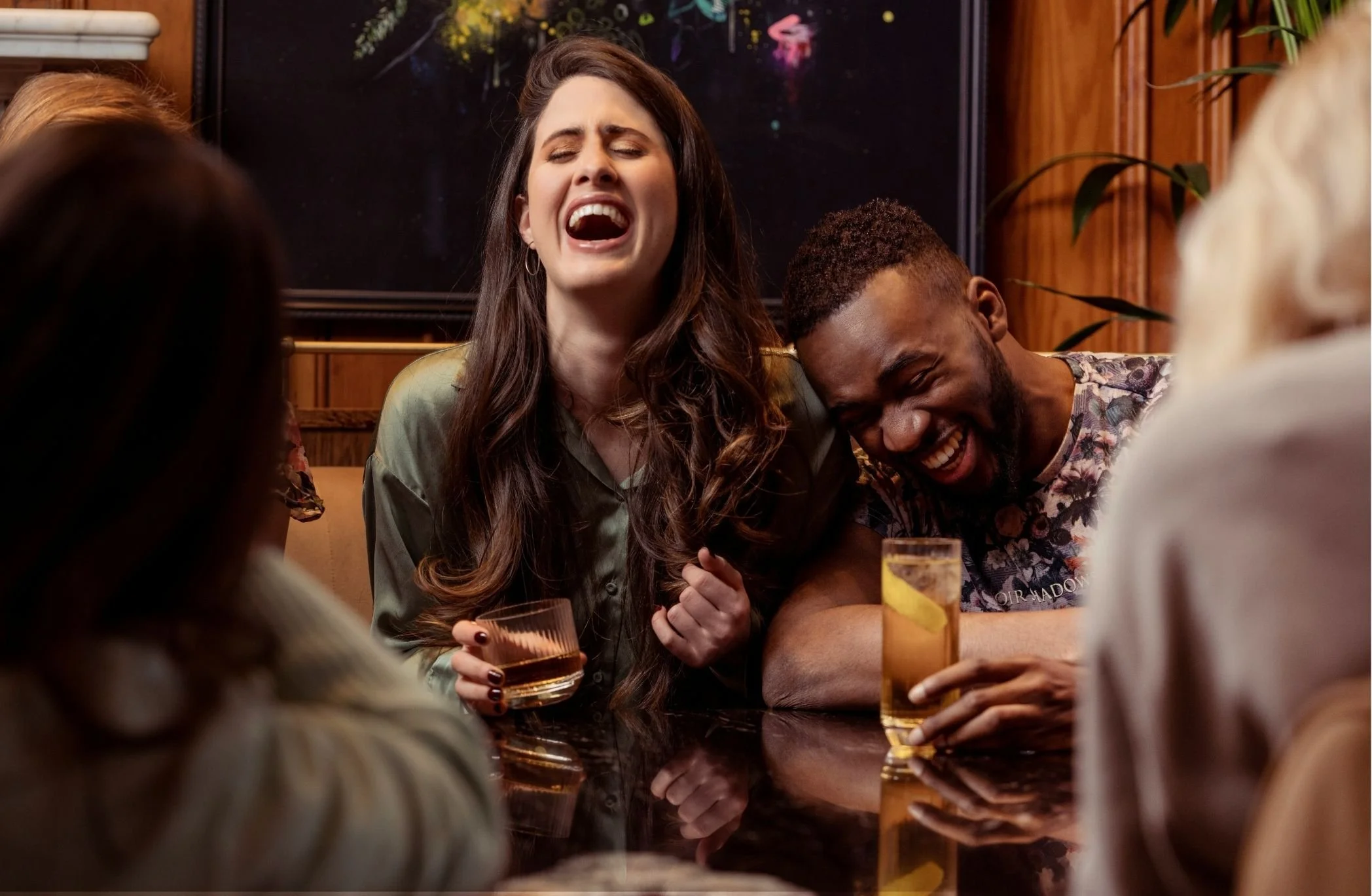 A group of friends at a bar, laughing and enjoying drinks, with a woman singing or laughing passionately.