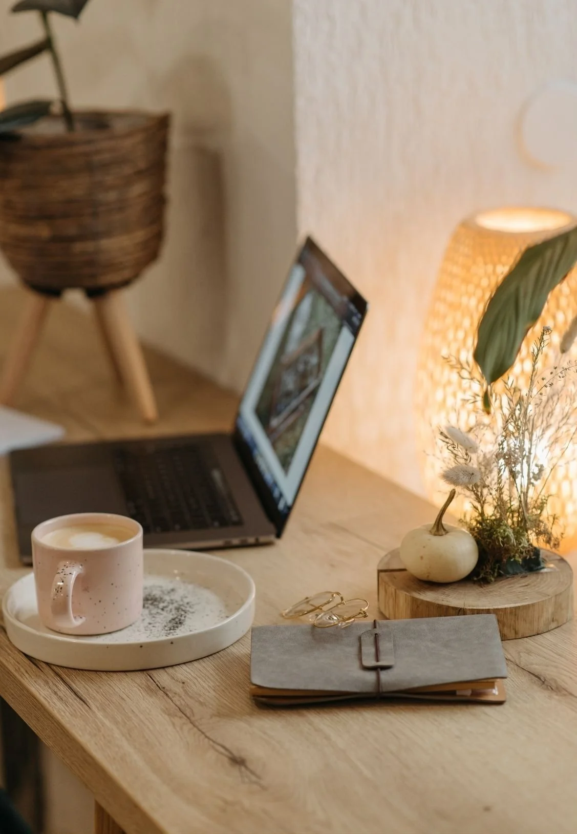 A cozy workspace featuring an open laptop, a pink coffee mug on a white tray, a small notebook with a leather strap, gold earrings, a decorative wooden tray with a gourd-shaped vase and dried flowers, and a warm table lamp, all on a wooden surface.