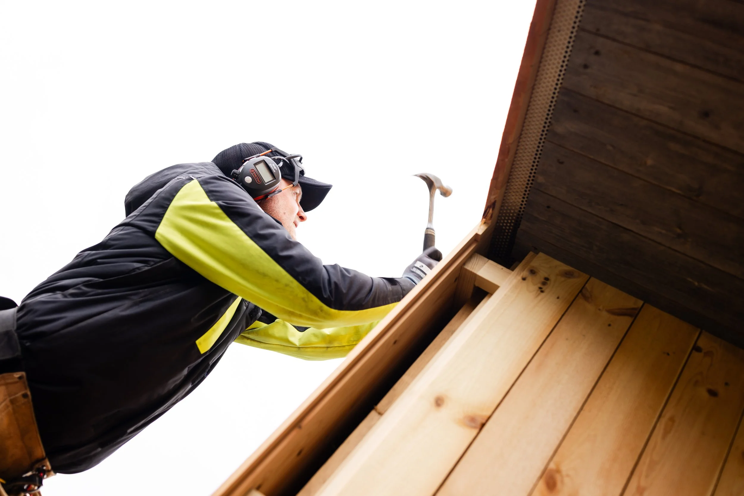 En mann som bruker hørselsvern og arbeidsklær, hamrer på en trevegg under takskifting.