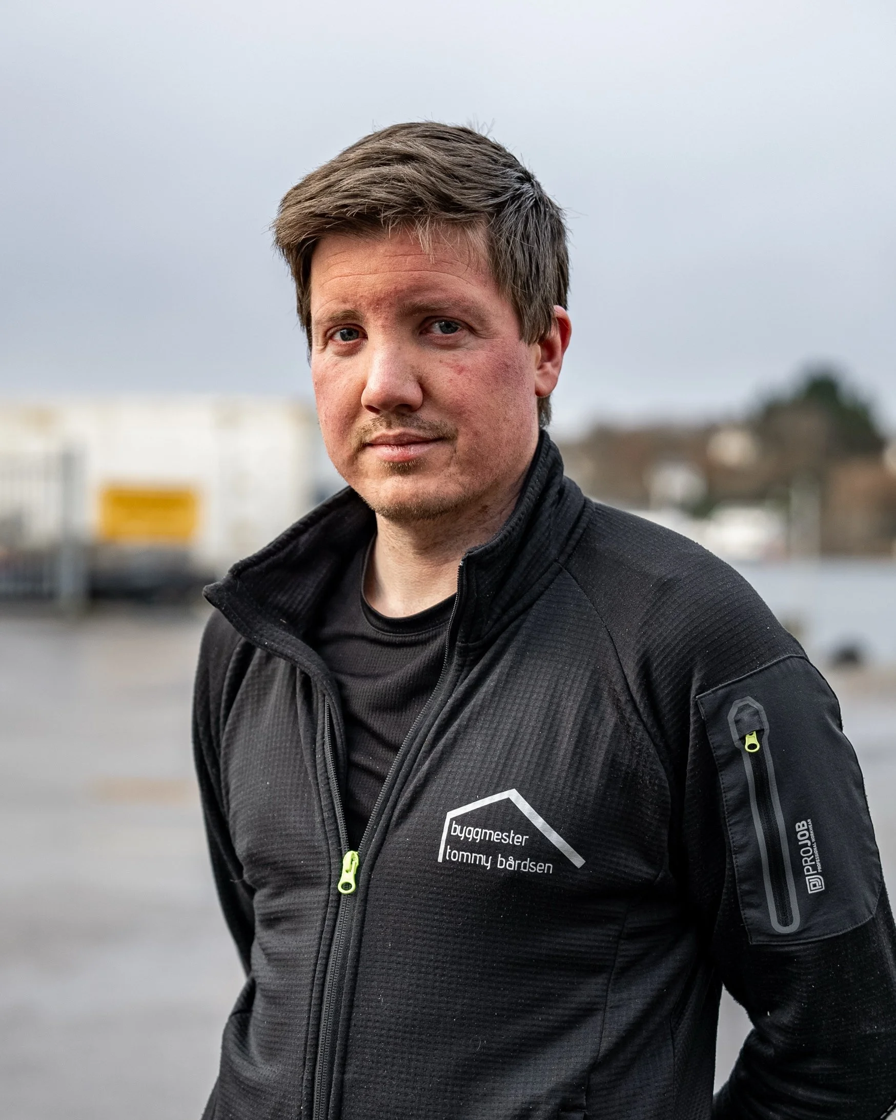 En mann som står utendørs, iført svart ytterjakke med logo og tekst, mot en bakgrunn av bygninger og en grå himmel.