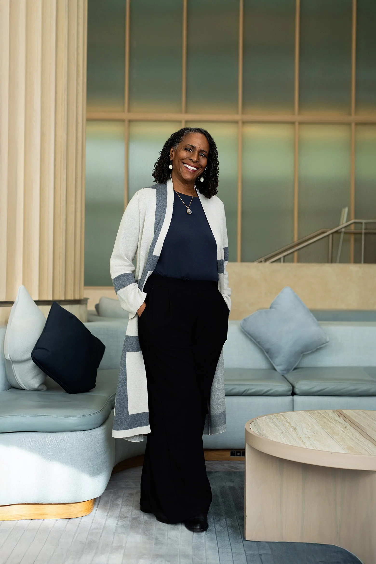 Woman smiling in a modern lounge area with light-colored seating, wooden accents, and a large window backdrop.