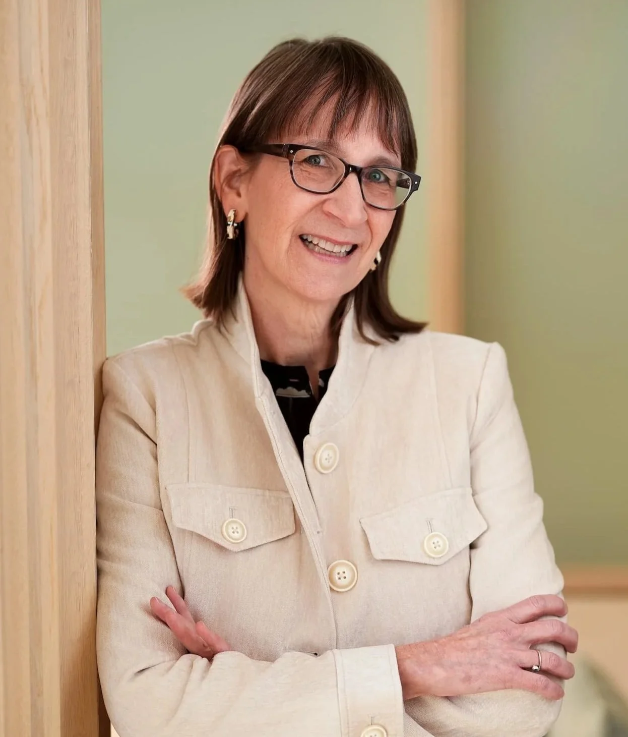 A woman with brown hair, glasses, and earrings standing with arms crossed, smiling, wearing a cream-colored blazer with large buttons, indoors with wooden and green background.