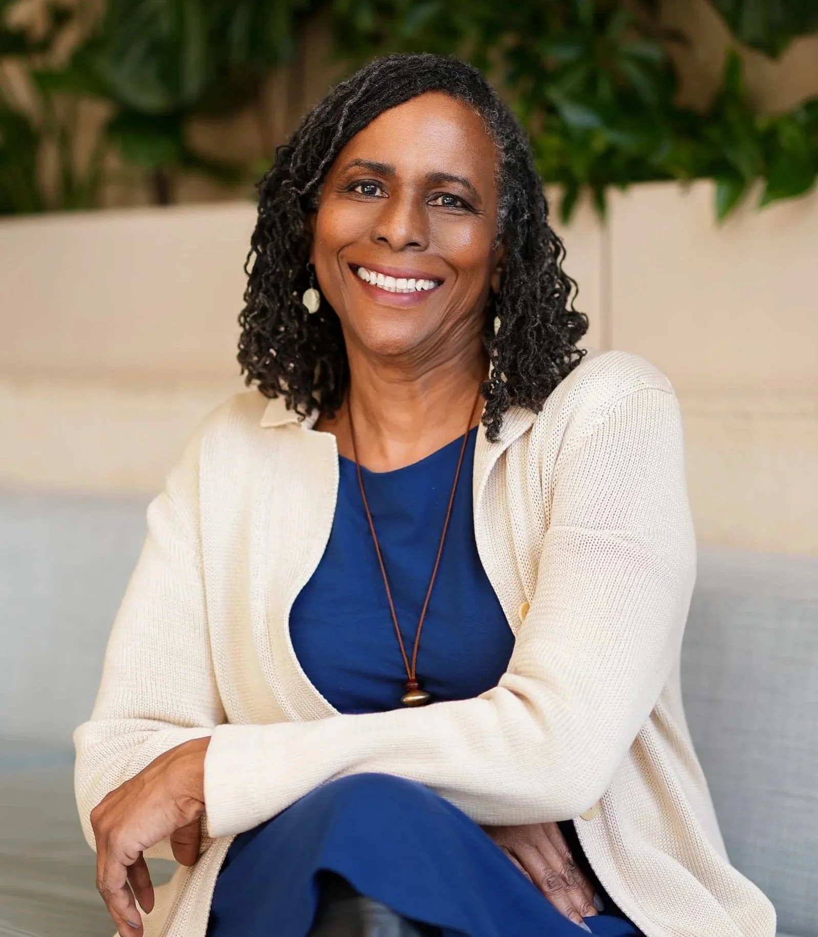 Smiling woman with curly black hair, wearing a cream-colored cardigan and blue shirt, sitting with arms crossed, in front of a background with green plants.