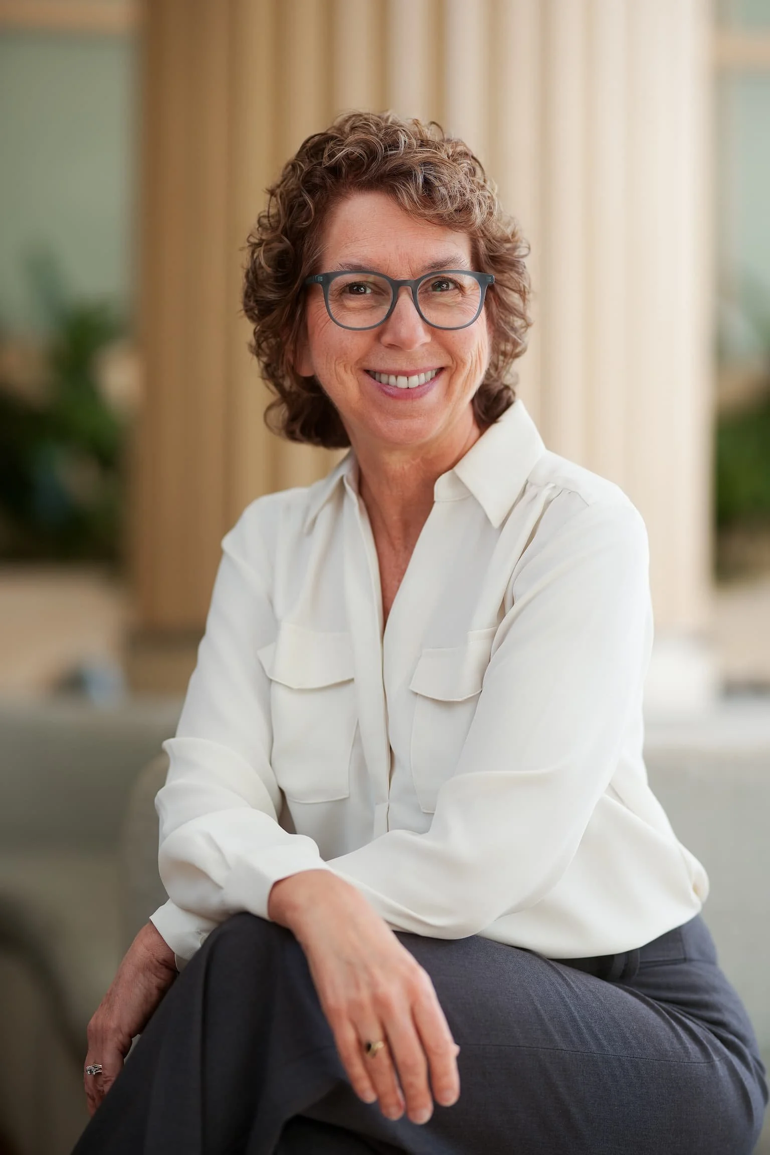 A woman with curly hair, glasses, and a white blouse sitting close to a person in dark clothing with their hand on her knee, both smiling indoors with beige curtains in the background.