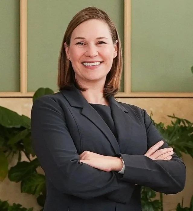 A woman with shoulder-length brown hair, smiling, wearing a dark blazer, with crossed arms, standing in front of a green wall and plants.