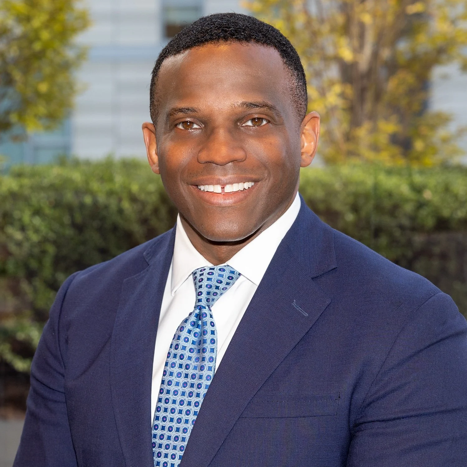 Smiling man wearing a blue suit jacket, white dress shirt, and blue, sitting in an office environment.