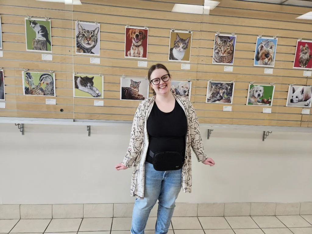 Young woman smiling at camera standing in front of wall display of framed cat and dog photographs at an art exhibit.