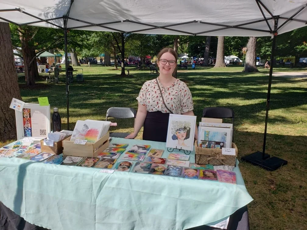 A woman standing behind a table at an outdoor art and craft fair, with various artwork, books, and stationery on display, under a canopy in a park with trees and other vendor tents in the background.