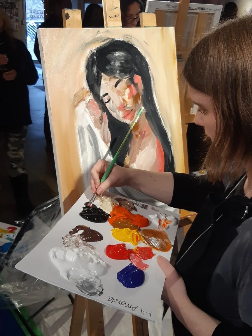 A woman painting an abstract portrait of a woman with black hair and pink highlights on a canvas. She is using a paintbrush and palette with various colors including white, black, red, yellow, brown, orange, and blue. The scene is inside an art studio with other people in the background.
