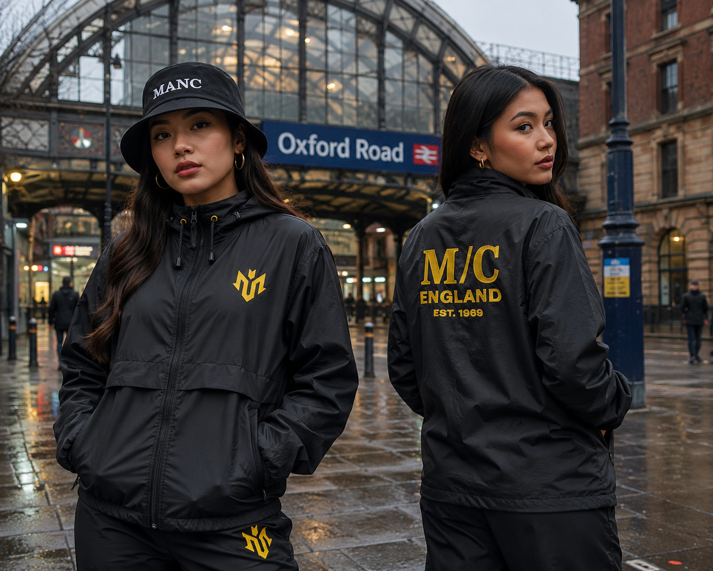 Two women wearing black jackets with yellow logos, standing in front of Oxford Road train station in Manchester, England, on a rainy day. One woman faces forward wearing a black hat, and the other is turned showing her back, displaying the text 'M/C England Est. 1969' on her jacket.