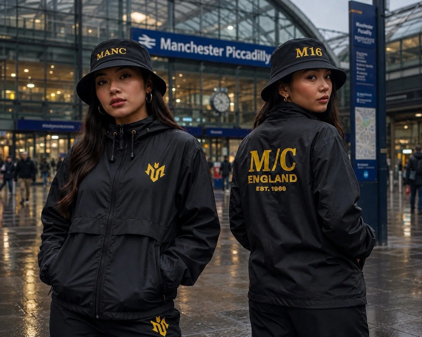 Two women wearing black jackets and hats with gold letters, standing outside Manchester Piccadilly train station in England, with a glass building and station sign in the background.