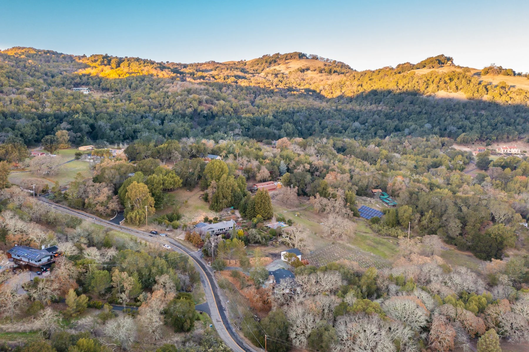 Aerial view of a rural landscape with trees, houses, a winding road, and mountains in the background during daytime.