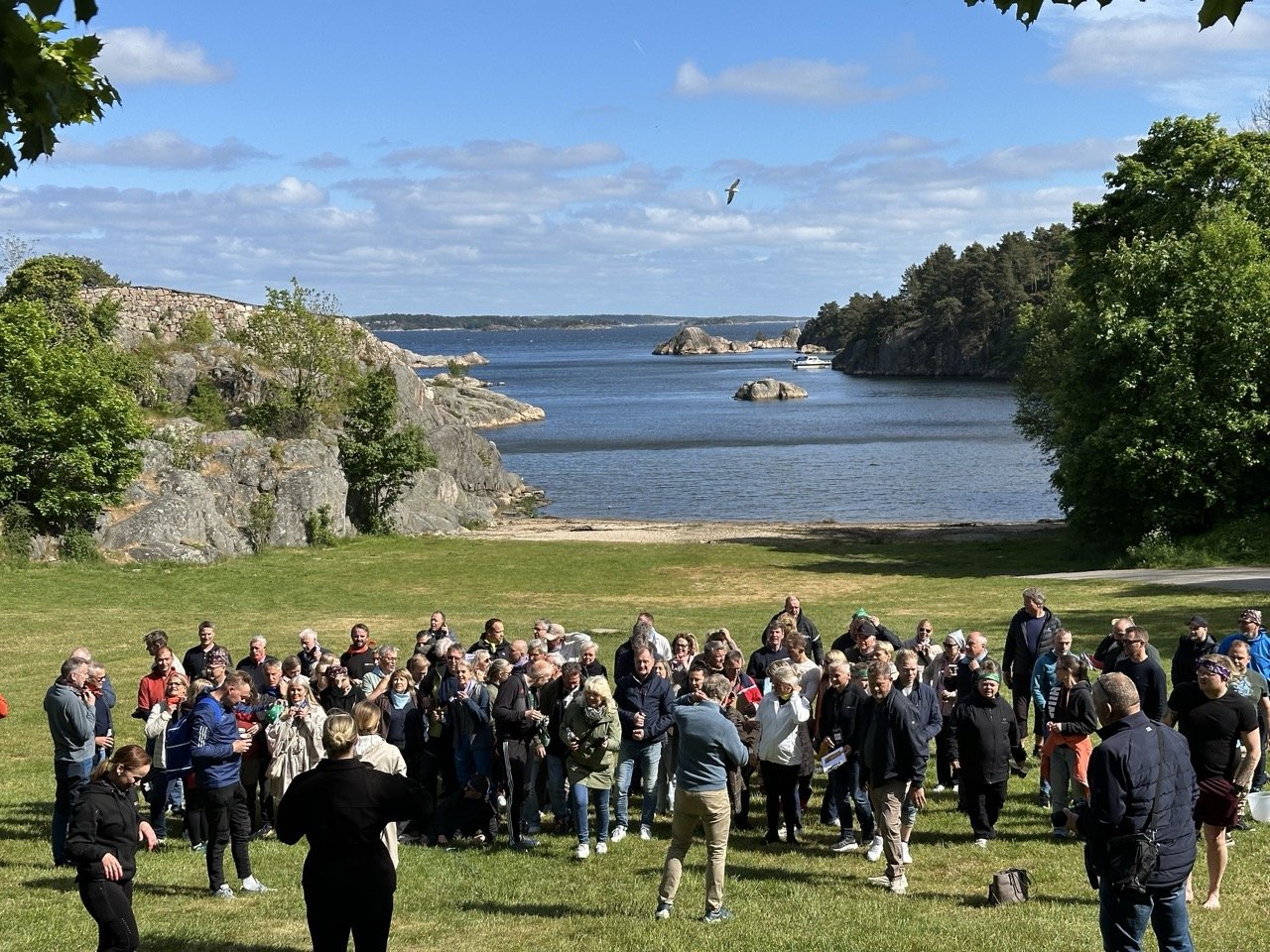 En gruppe mennesker samles på en grønn plener foran en innsjø omgitt av steinete klipper og trær under en blå himmel med skyer, med en seilflyver i luften.