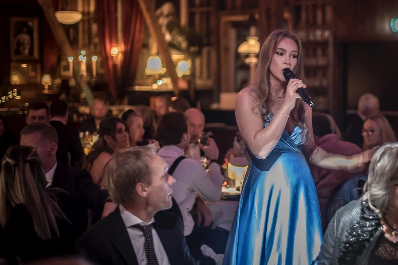En kvinne i en blå kjole synger med mikrofon på en kveld i et restaurant- eller festlokale med mange mennesker rundt henne.